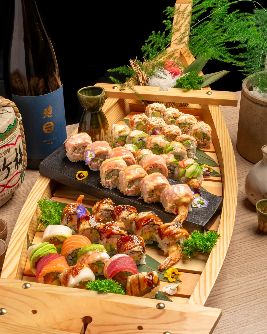 Dřevěná loď plná sushi rolek a nigiri vedle lahve saké